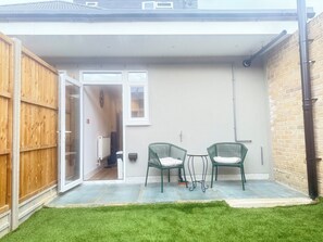 Eine Terrasse mit zwei grünen Stühlen, einem kleinen Tisch, einem Holzzaun und einem Hauseingang.