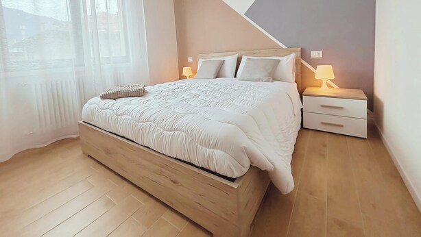 A bedroom with a wooden bed, white bedding, a bedside lamp, a nightstand, and a window with sheer curtains. - The Lake House Cernobbio -  Village Center, Walk to Lake (Cernobbio)