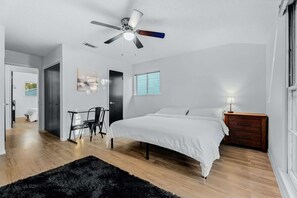 A bedroom with a bed, a desk, a chair, a ceiling fan, and a window.