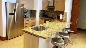 Una cocina moderna con refrigerador de acero inoxidable, encimeras de granito, horno integrado y un lavamanos. Hay tres taburetes de bar y armarios de madera.