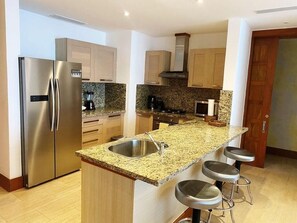 Eine moderne Küche mit einem Edelstahlkühlschrank, Granitarbeitsplatten, einem eingebauten Ofen und einem Spülbecken. Es gibt drei Barhocker und Holzschränke.