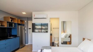 A compact hotel room with a kitchenette, a bed, a mirror, and a view of the city. - Studio Just 500m From Vila Germanica Pin1905 (Blumenau)
