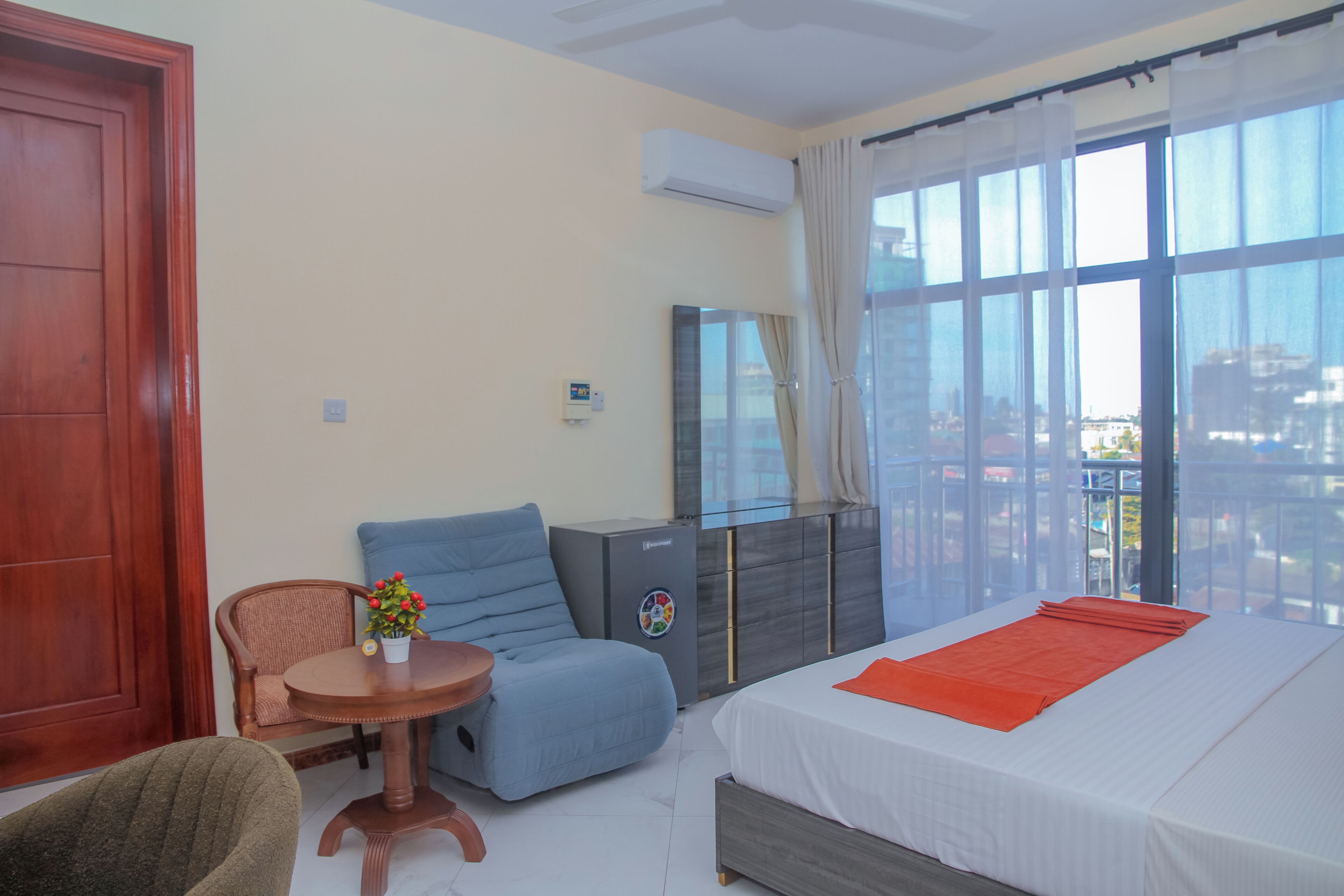 Ein Hotelzimmer mit Bett, Sessel, kleinem Tisch mit Topfpflanze, Kleiderschrank und großem Fenster mit Vorhängen.