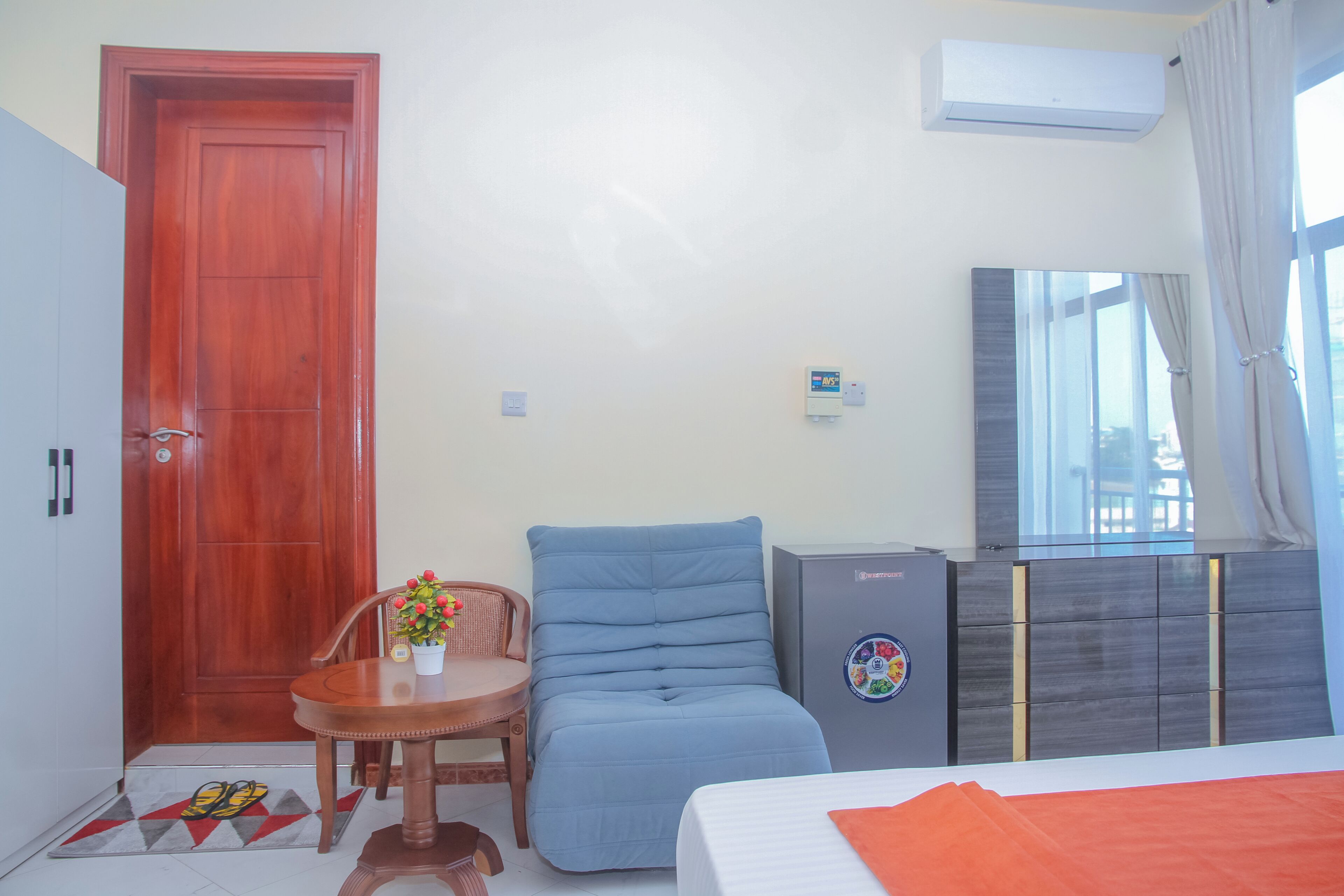 A room with a wooden door, a blue chair, a small table with a potted plant, a bed with an orange blanket, and a grey cabinet.