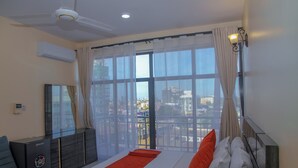 Un dormitorio con cama, ventilador de techo, aire acondicionado, cómoda y vistas a la ciudad a través de amplias ventanas.
