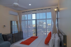 A bedroom with a bed, a ceiling fan, an air conditioning unit, a dresser, and a view of the city through large windows. - ANDREW VIEW HOTEL (Dar es Salaam)