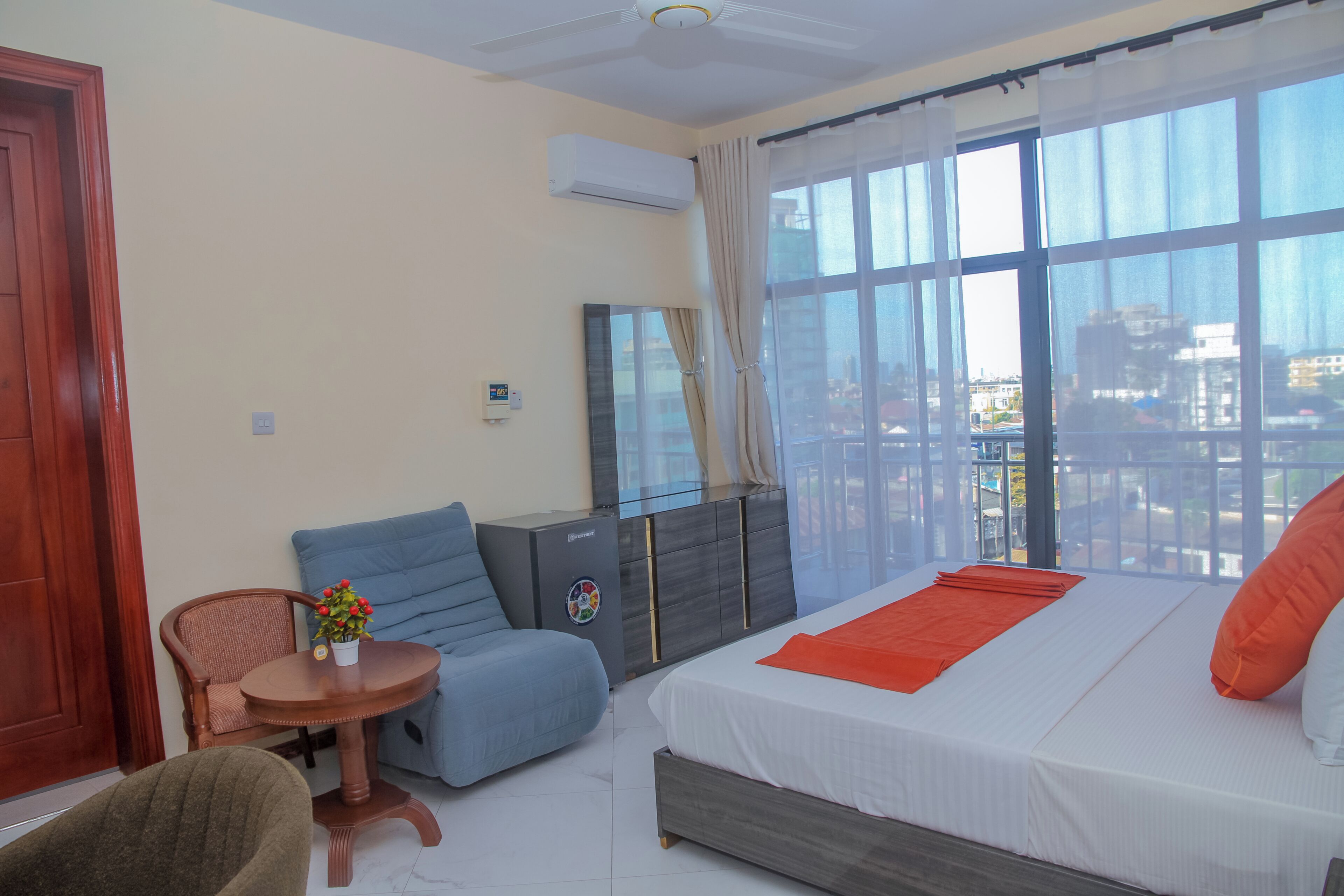 Une chambre d’hôtel avec un lit, une chaise, une petite table avec une plante, une armoire et une fenêtre donnant sur des immeubles.