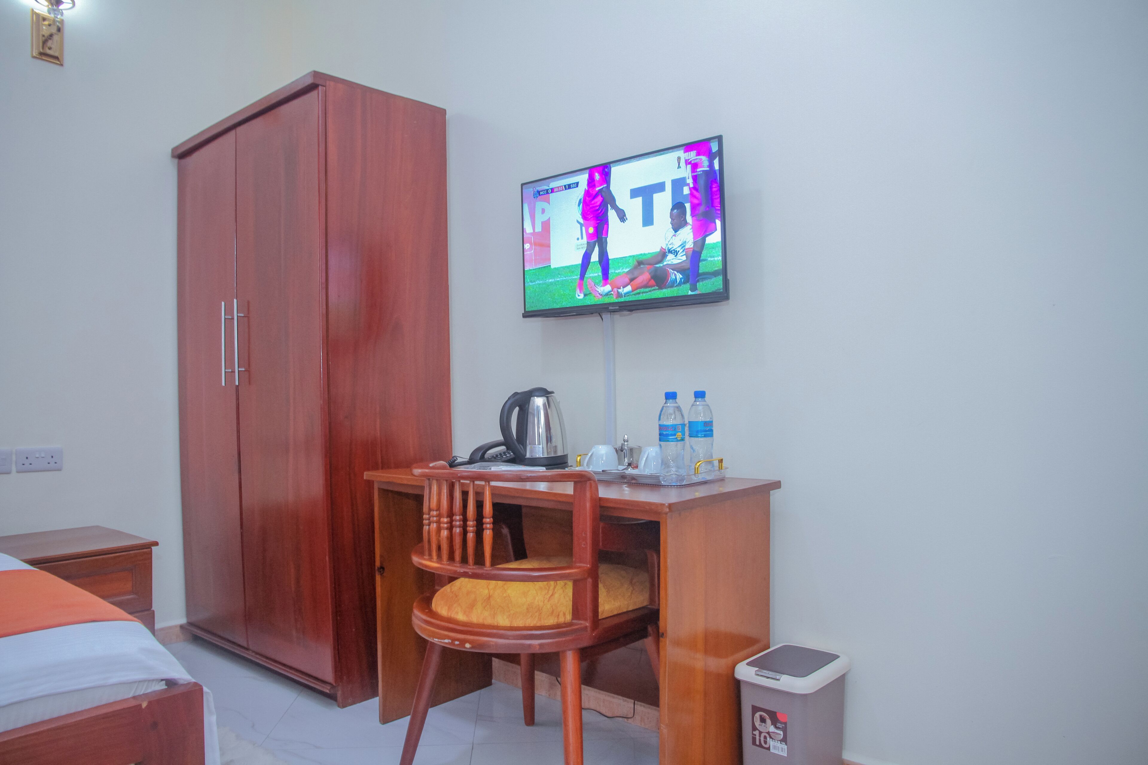 Una habitación con un armario de madera, un escritorio con una silla, un televisor montado en la pared y un cubo de basura.