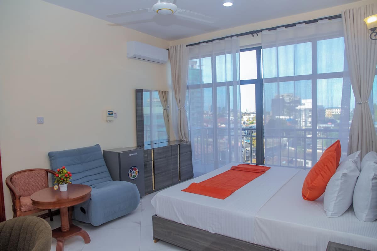 Habitación de hotel con cama, una silla, una mesita con un florero, cómoda y vistas a través de amplios ventanales.