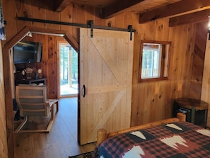 Ein Interieur einer Holzhütte mit einem Bett, einem Schreibtisch, einem Stuhl, einem Fenster und einer Tür.