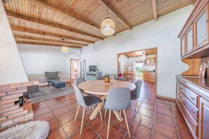 Ein geräumiger Wohnbereich mit einem runden Holztisch, grauen Stühlen, einem Kamin aus Ziegelsteinen und einer hohen Holzbalkendecke.