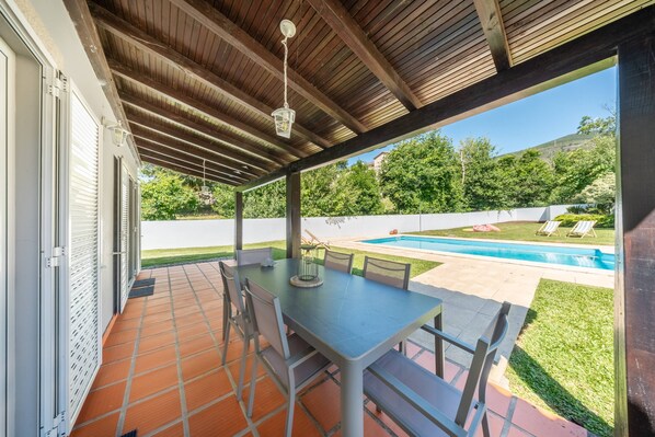 Un patio couvert avec une table et des chaises, un luminaire suspendu et une vue sur une piscine et la verdure.