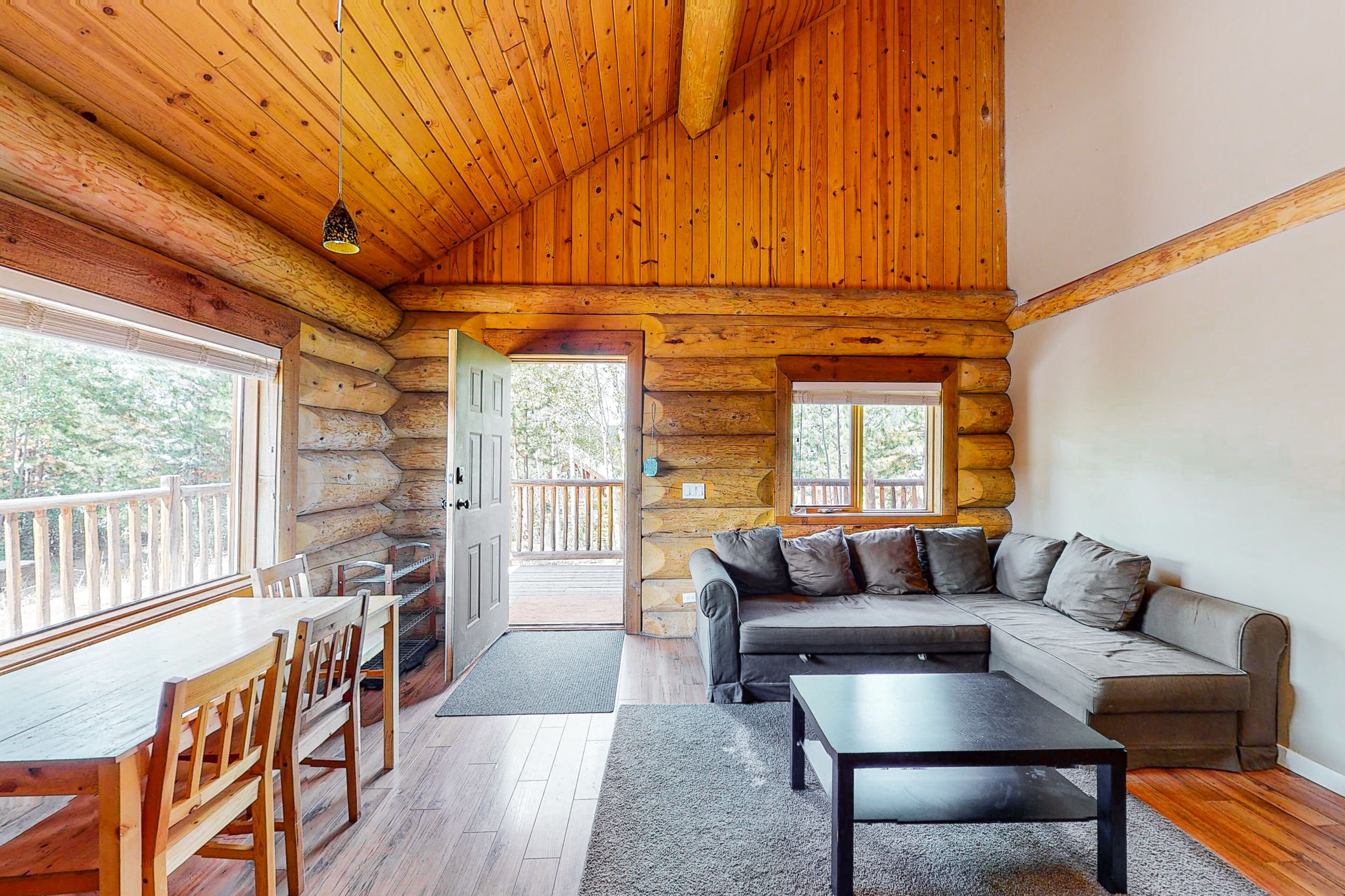 Interior de una cabaña de madera con comedor, sofá y vista por la puerta.
