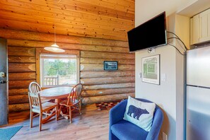 Interior de una cabaña de troncos con comedor, un sofá azul, televisor de pantalla plana y refrigerador.