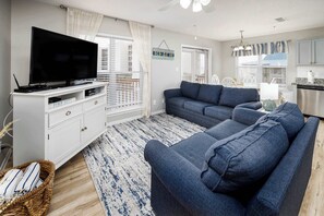 A living room with a flat-screen TV, a sofa, a white entertainment unit, and a kitchen area in the background. - Sunset Cottages 5C 🏖️Free Beach Setup (Fort Walton Beach)