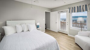 Ein Schlafzimmer mit einem großen Bett, einem Nachttisch, einer Lampe, einem Fenster mit Jalousien und einer Tür zu einem weiteren Raum.