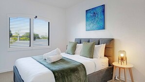 A bedroom with a bed, pillows, a blanket, a painting, a window with blinds, and a bedside lamp.