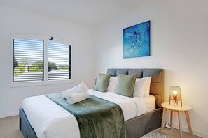 A bedroom with a bed, pillows, a blanket, a painting, a window with blinds, and a bedside lamp. - Ponsonby 4BR Townhouse w Parking | Walk to Shops (Auckland Central Suburbs)