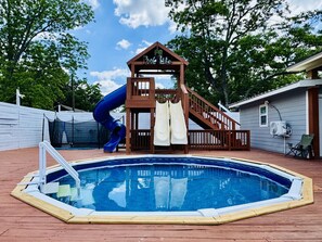 Ein Schwimmbecken mit Holzterrasse, einer Rutsche und einer Spielanlage aus Holz.