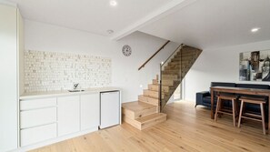 Eine moderne Küche mit weißen Schränken, einer gefliesten Spritzschutzwand und einer Holztreppe, die in einen Essbereich mit einem schwarzen Sofa und Holztischen führt.