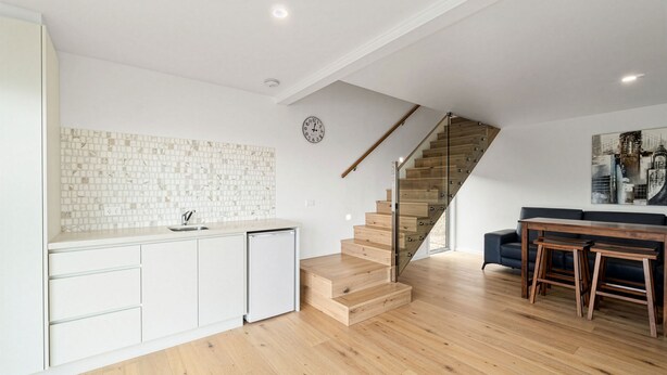 Une cuisine moderne avec des meubles blancs et un pare-feu en carreaux, un escalier en bois et un coin repas avec un canapé noir et une table en bois.