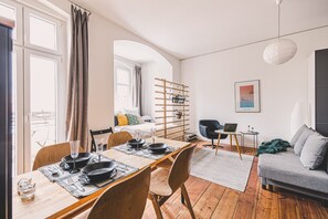 A modern living room with a dining area, a grey sofa, a wooden dining table set for two, and a laptop on a side table. - Diente | Studio | Poznań Jeżyce | Balkon & Kamienica (Poznań)
