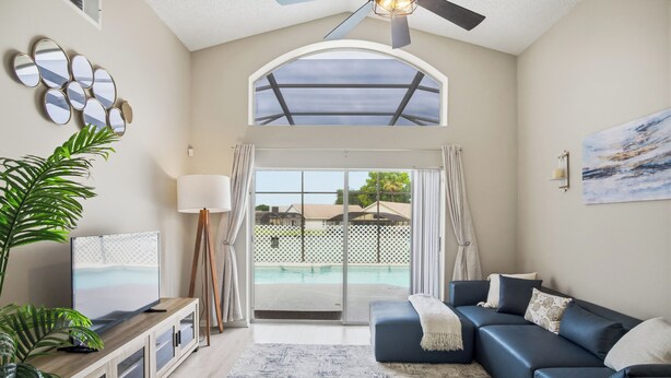 A living room with a TV on a wooden cabinet, a blue sofa, a large plant, and a view of a swimming pool through glass doors. - Spacious 4BRFamily Retreat-Pool Minutes to Disney (Davenport)