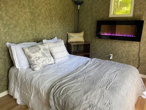 Ein Bett mit grauer Tagesdecke, eine Nachttischlampe, ein kleines Regal mit einem Kopfkissen und ein an der Wand montierter Fernseher.