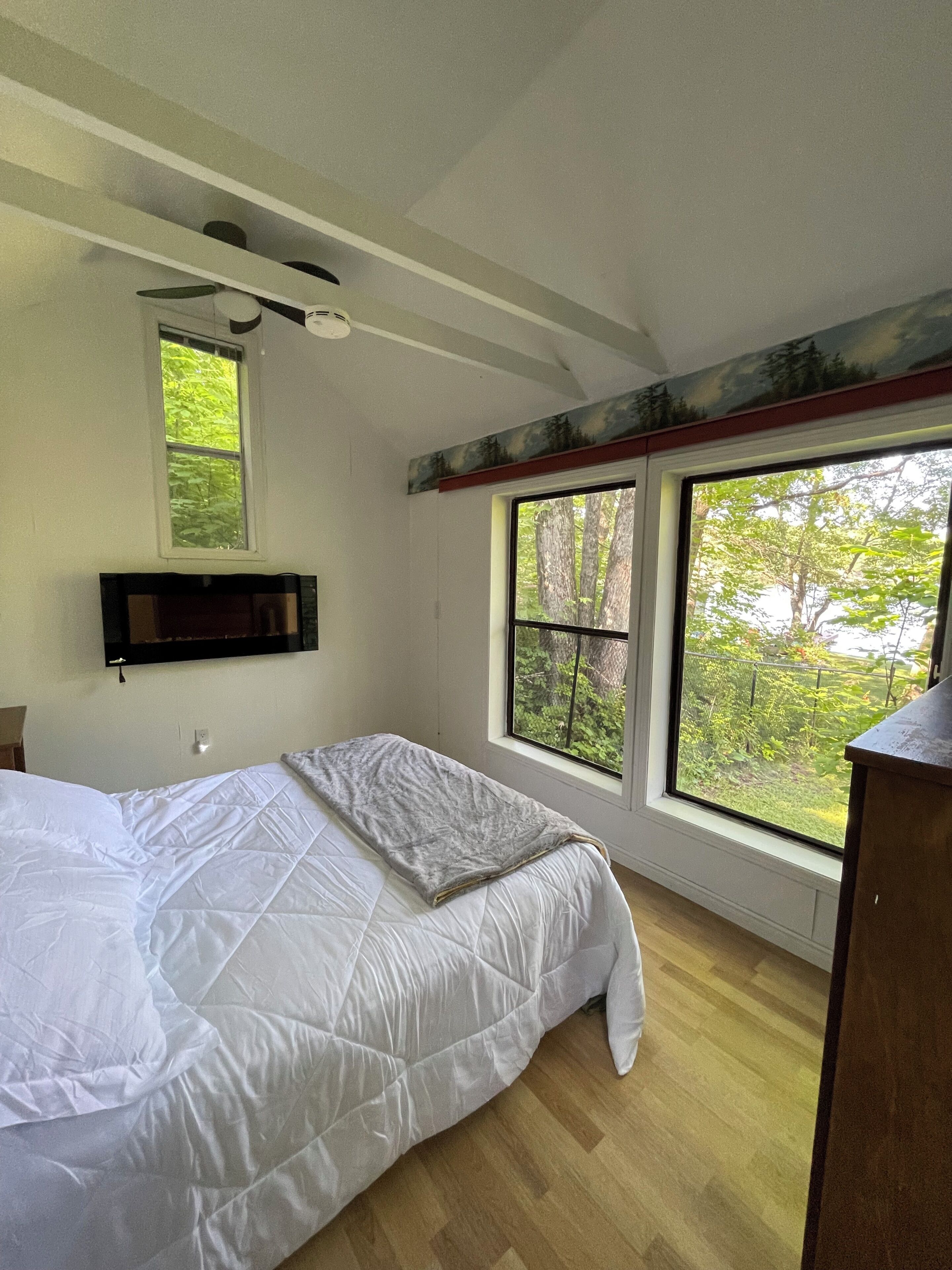 A bedroom with a bed, a mounted TV, a window with a view of trees, and a ceiling fan.