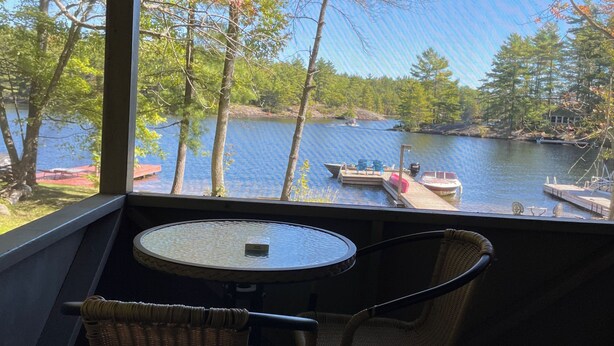 Un área de estar al aire libre, techada, con una mesa y sillas, con vista a un lago con barcos y árboles.