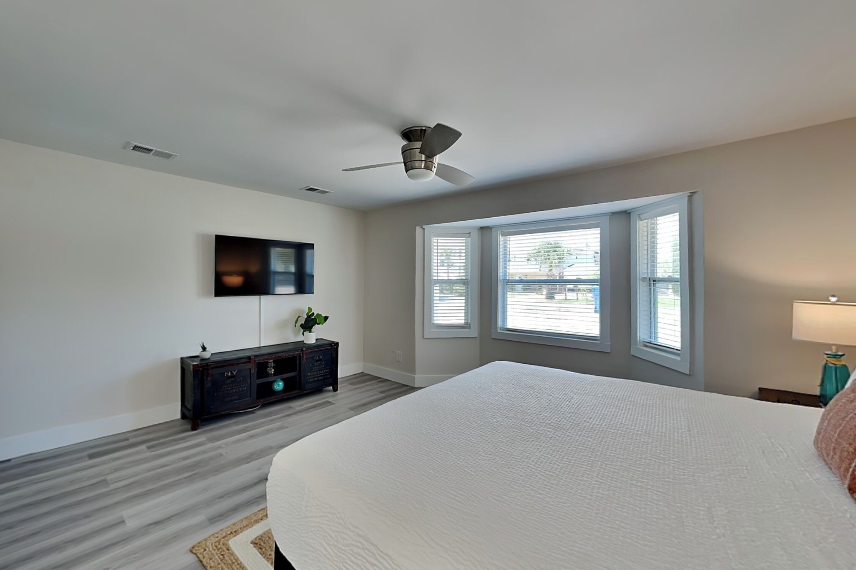 A bedroom with a bed, a ceiling fan, a TV, a window with blinds, and a nightstand with a lamp.