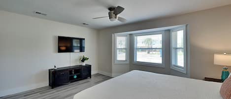 A bedroom with a bed, a ceiling fan, a TV, a window with blinds, and a nightstand with a lamp.