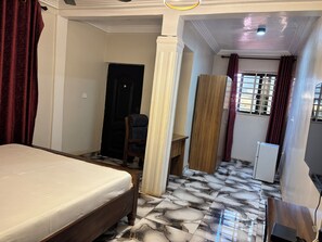 A hotel room with a bed, a desk, a chair, a wardrobe, and a refrigerator. - KELNESSA HOTEL (Obuasi)