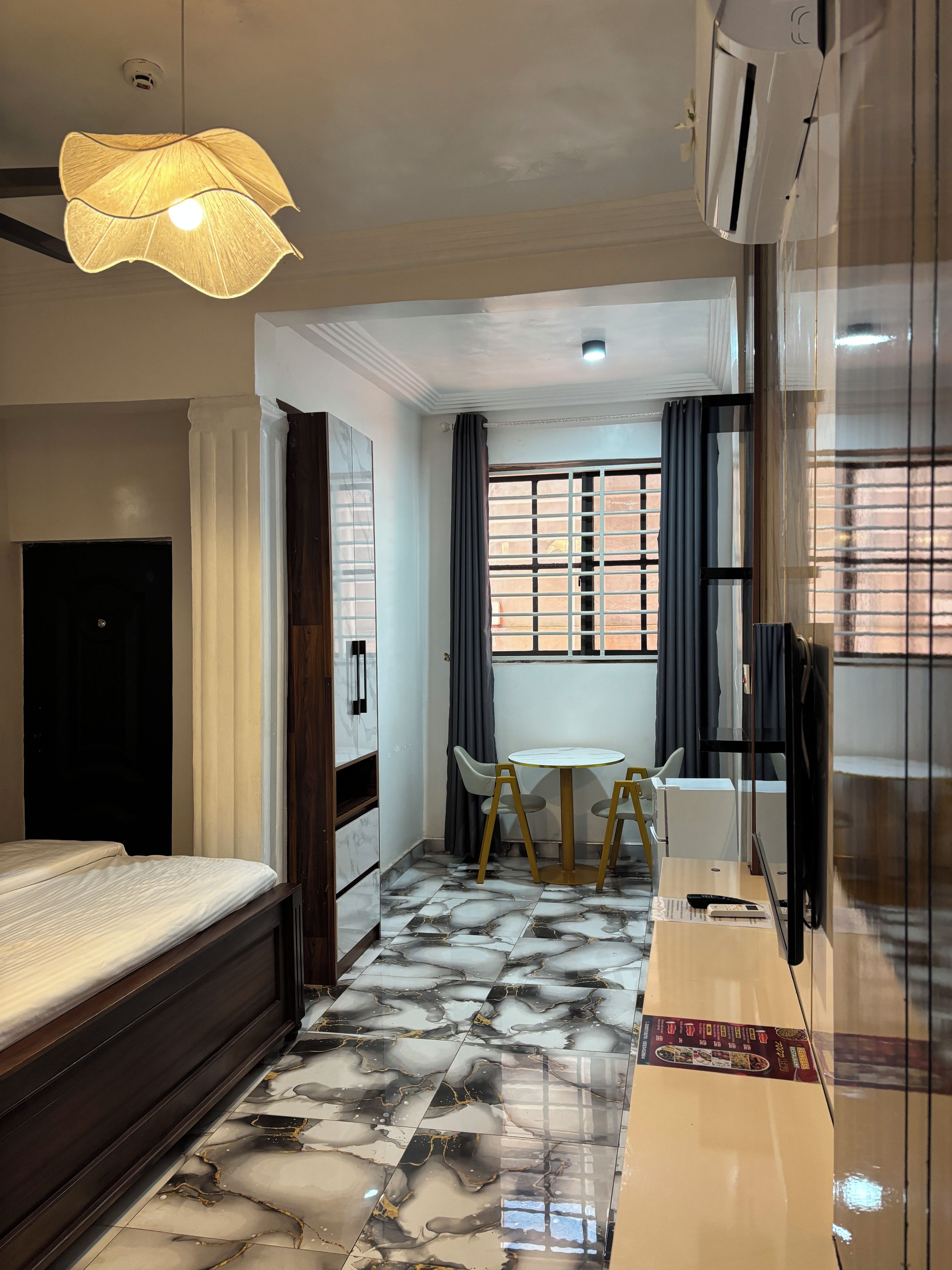 A hotel room with a bed, a small table with chairs, a window with curtains, and an air conditioning unit.