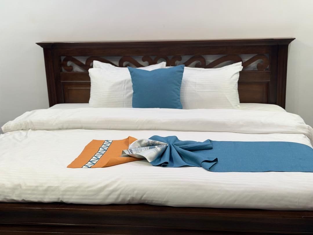 Een net opgemaakt bed met witte lakens, een blauw kussen en een oranje en blauwe deken.