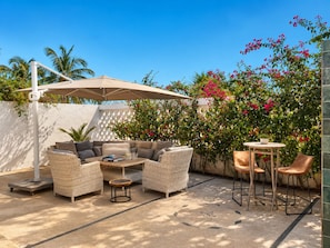 An outdoor patio with wicker furniture, a table, and chairs, surrounded by lush greenery and flowering plants.