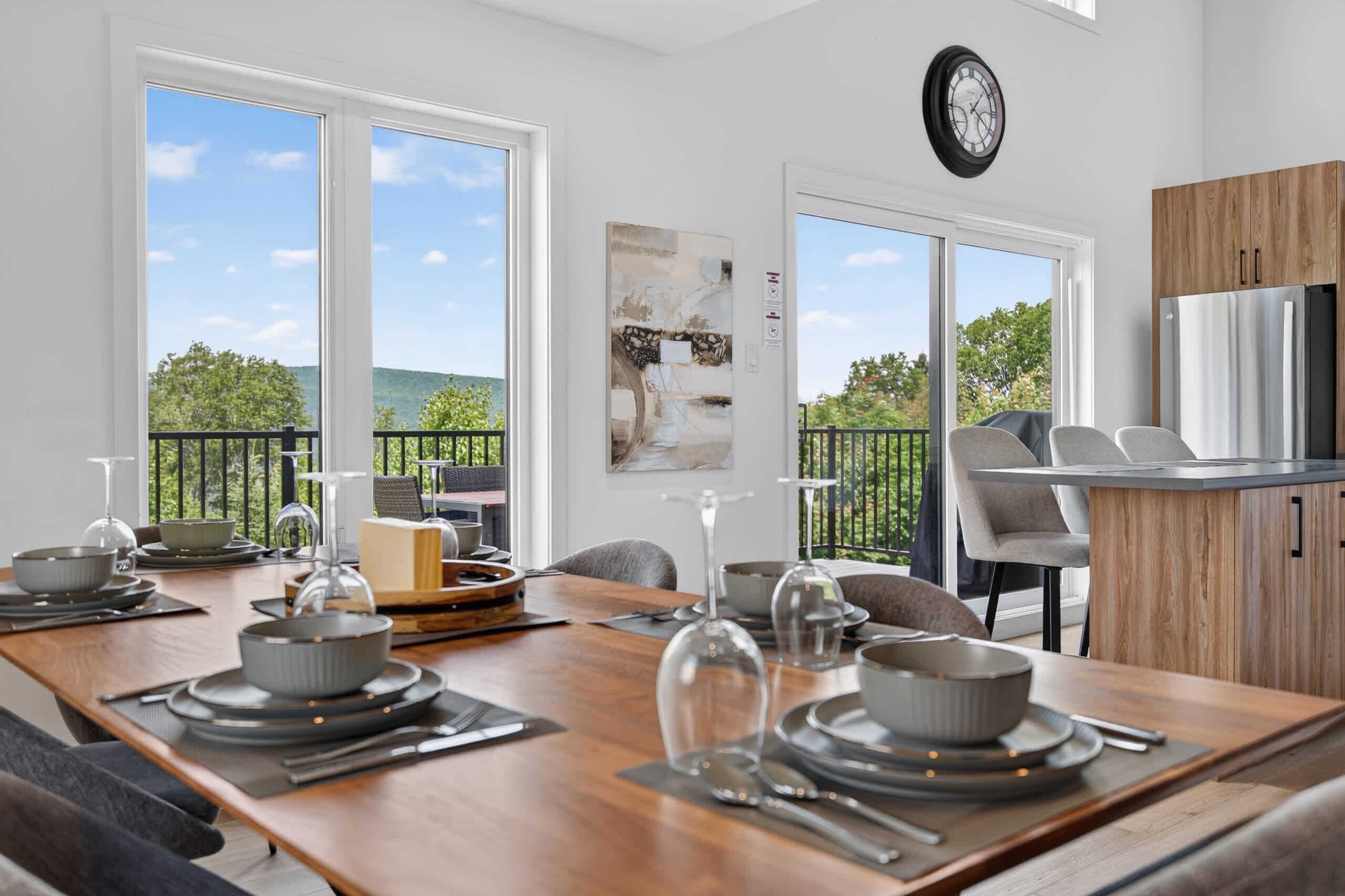 A dining area with a wooden table set for four, a kitchen with a refrigerator, and a balcony with a view of trees and hills.
