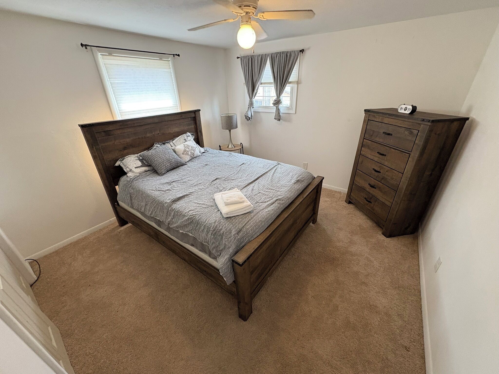 Ein Schlafzimmer mit einem hölzernen Bett, einem Nachttisch, einer Kommode, einem Deckenventilator und einem Fenster mit Vorhängen.
