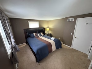 A bedroom with a bed, a nightstand, a lamp, a window with curtains, and a door.