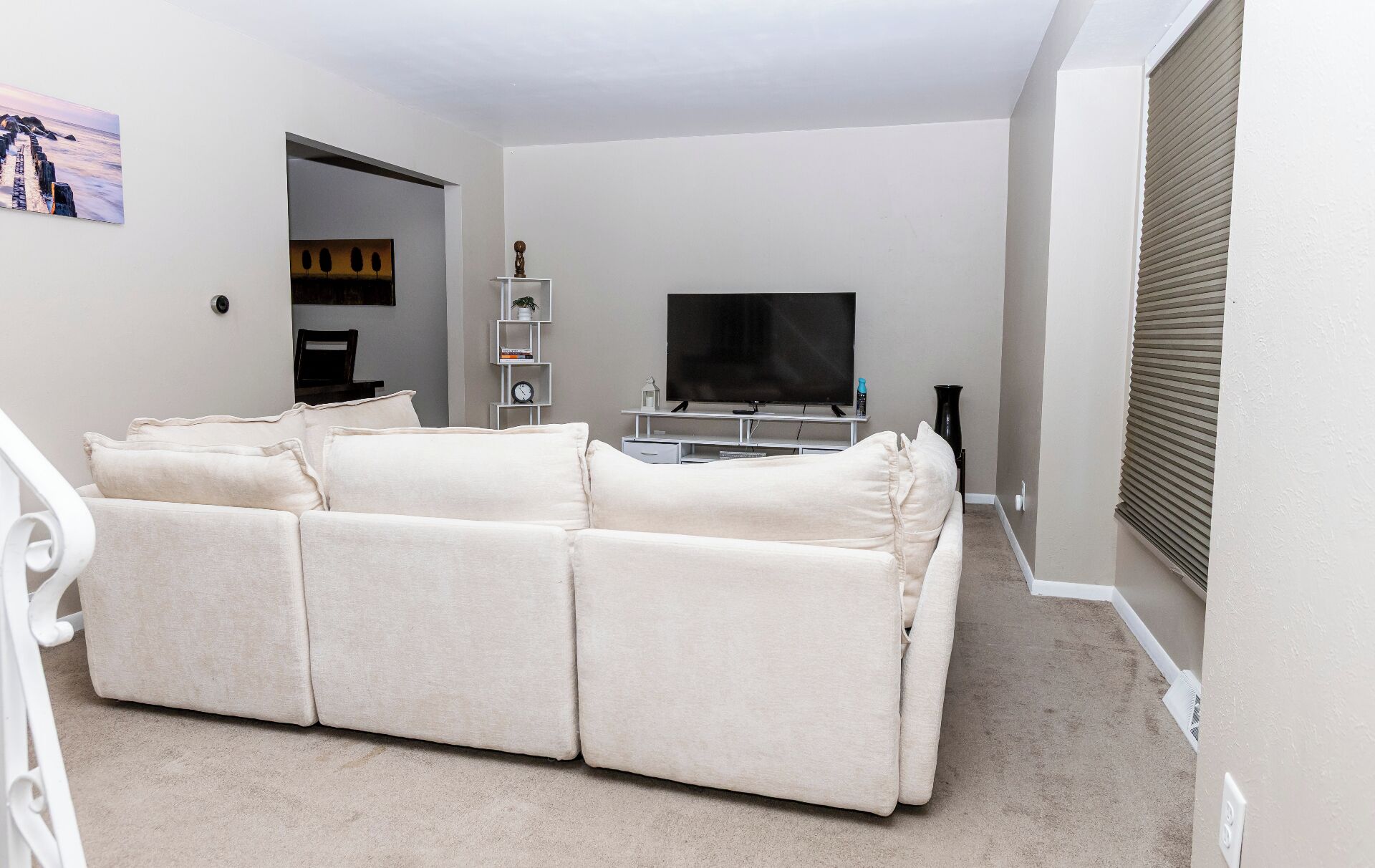 A living room with a beige sofa, a flat-screen TV, a small white shelf with books, and a painting on the wall.
