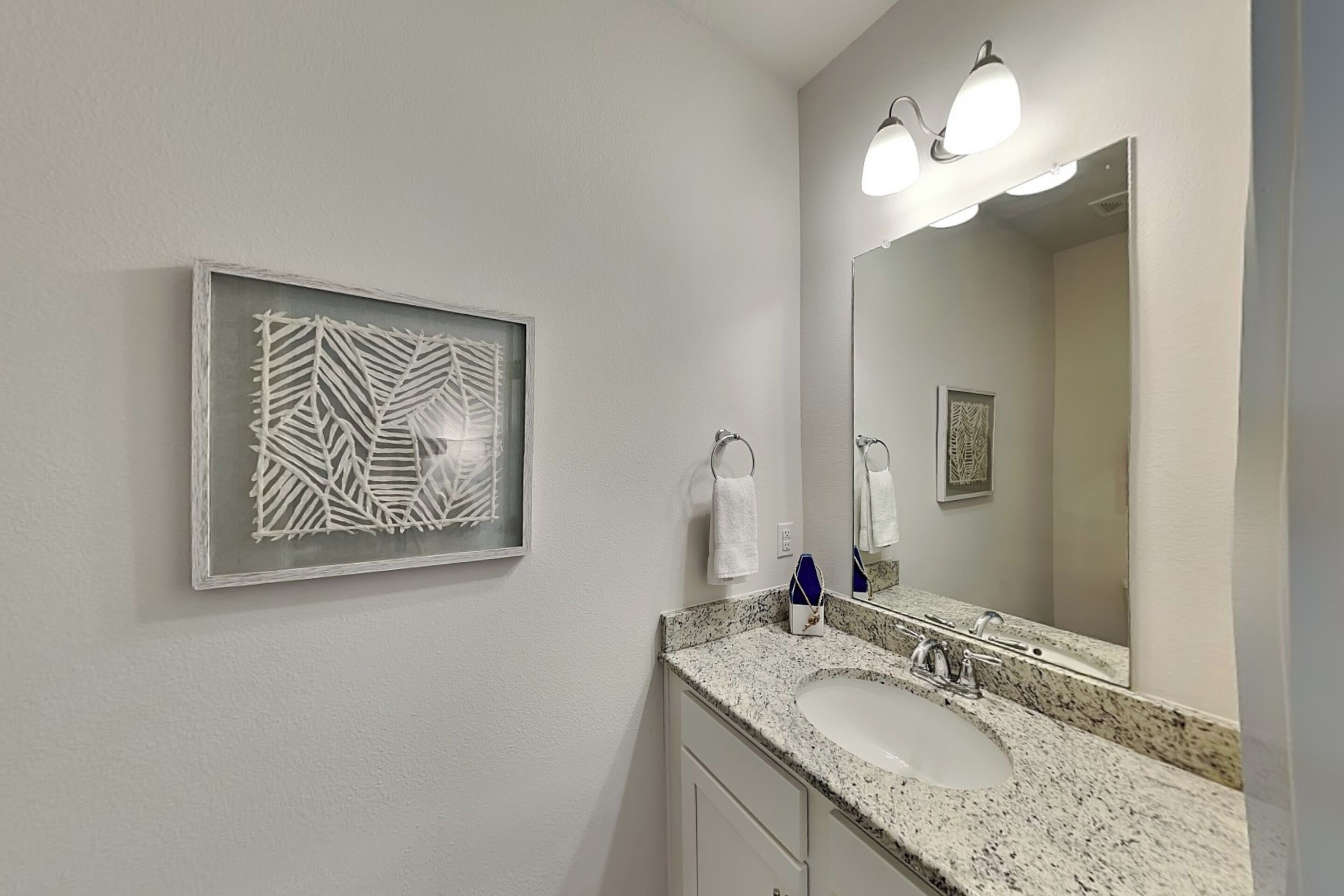 Un baño con encimera de granito, un lavamanos blanco, un espejo grande y una obra de arte abstracta enmarcada en la pared.