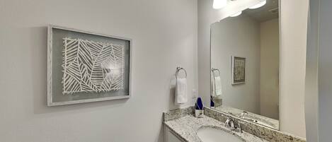 Un baño con encimera de granito, un lavamanos blanco, un espejo grande y una obra de arte abstracta enmarcada en la pared.