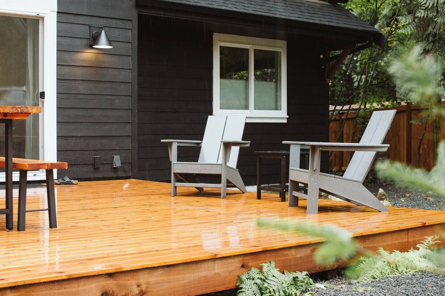 Um deck de madeira com duas cadeiras Adirondack, uma mesa e uma janela com persianas.