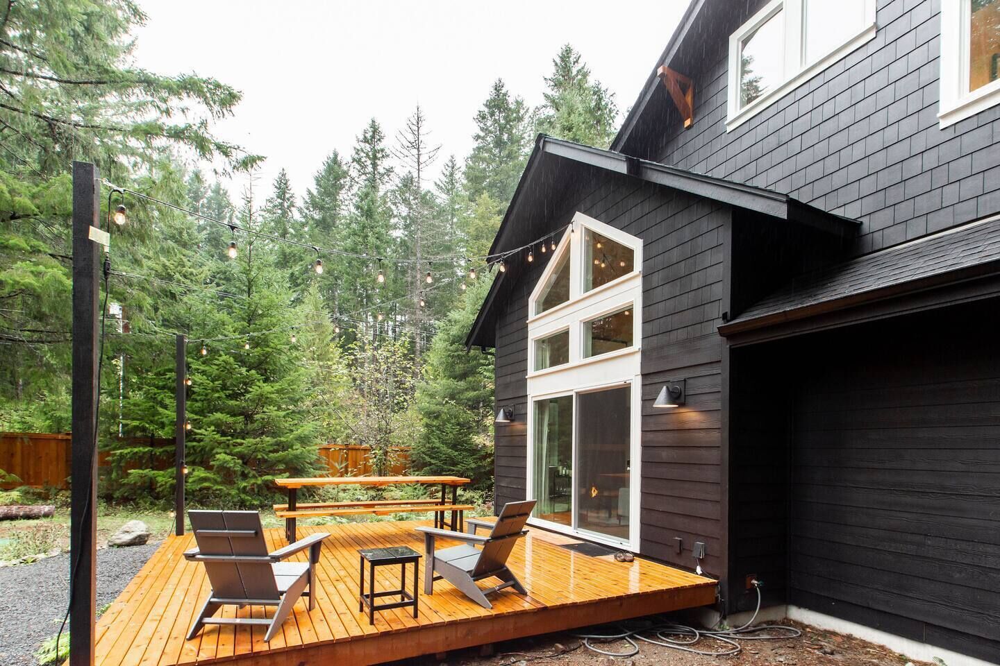 A modern house with a deck, outdoor seating, and a forest backdrop.