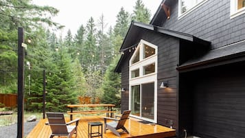 A modern house with a deck, outdoor seating, and a forest backdrop.