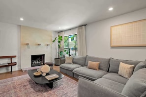 A modern living room with a grey sofa, a black coffee table, a fireplace, and a large window with curtains. - Laurel Canyon 5BR | Pizza Oven + Fireplaces | C&C (Los Angeles)