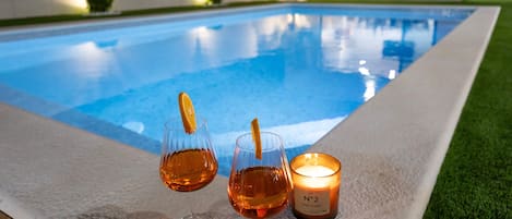 A poolside setting with two glasses of amber liquid, a lit candle, and a pool in the background.