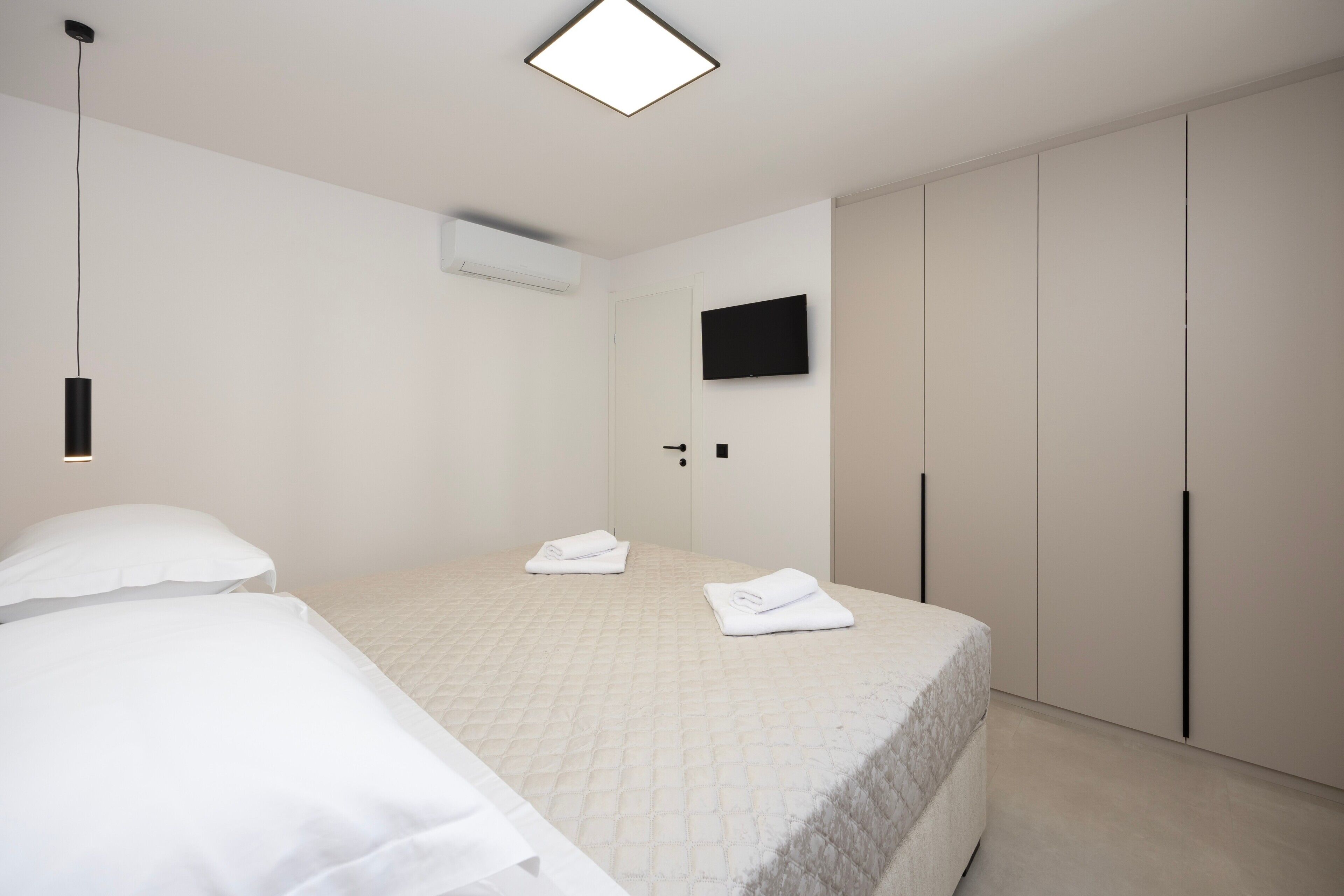 A bedroom with a bed, two pillows, a flat-screen TV, a wardrobe, and a ceiling light fixture.