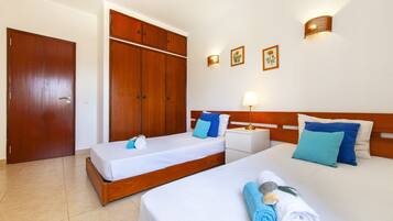 Quarto de hotel com duas camas de solteiro, um guarda-roupa, uma mesa de cabeceira com abajur e uma luminária na parede.