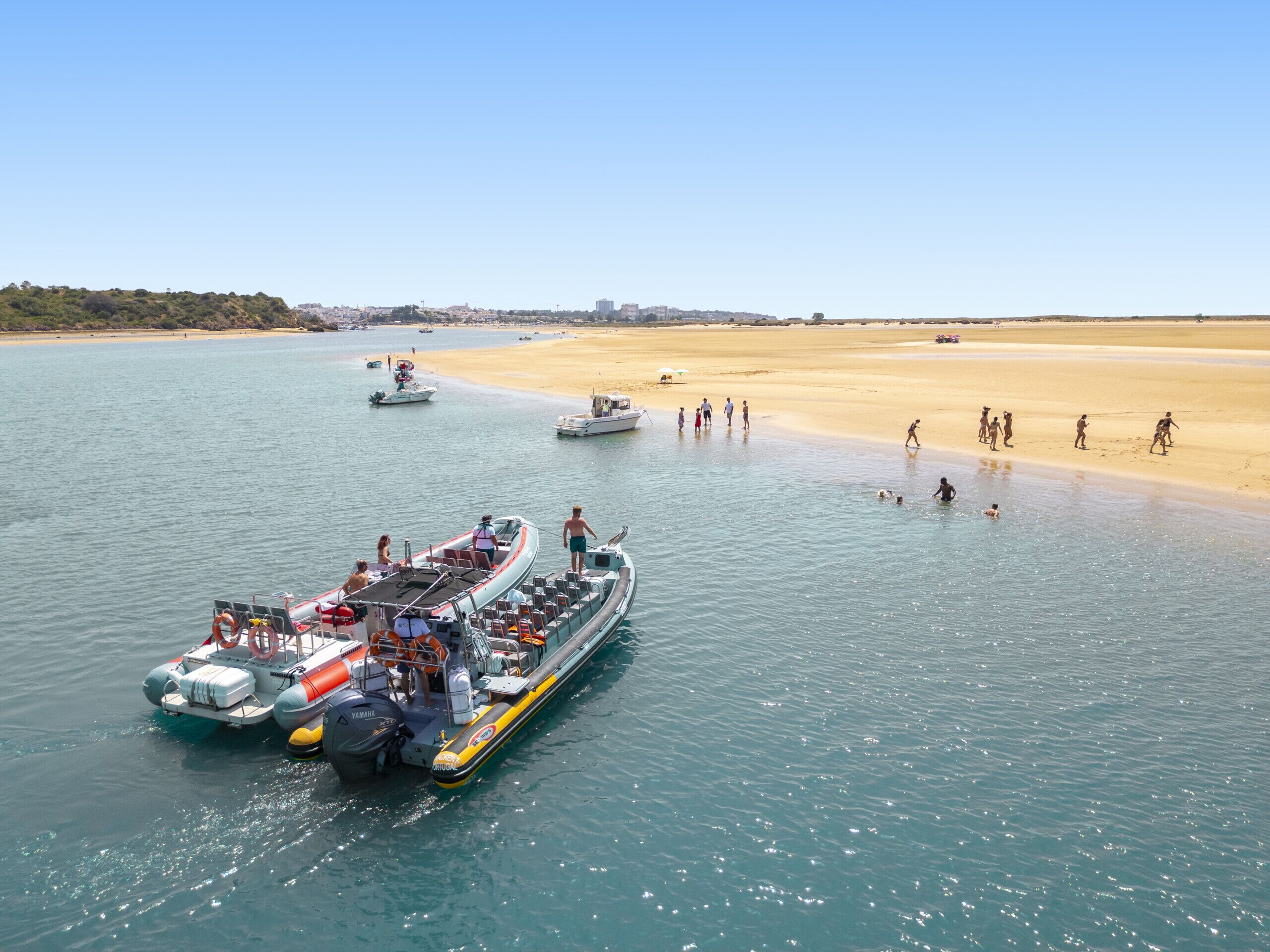 A ferry docked at a beach with people walking on the shore and boats in the water.